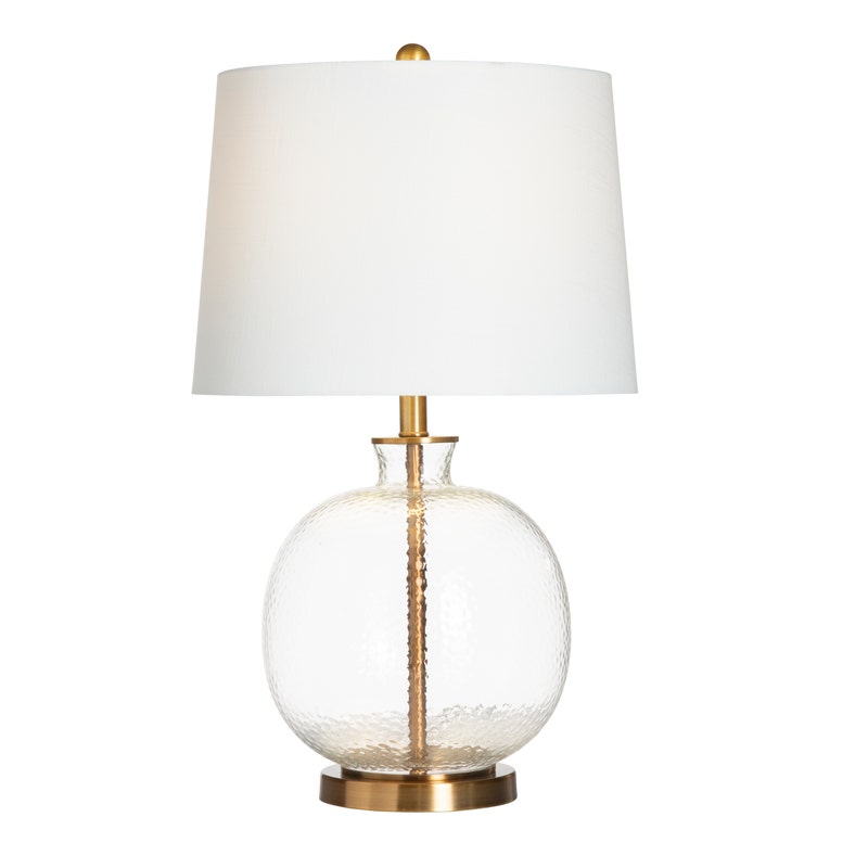 Table lamp with clear glass base, gold accents, and white shade on a white background