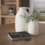 Two white ceramic vases with a plant on a wooden surface, with a book and decorative item in the foreground.
