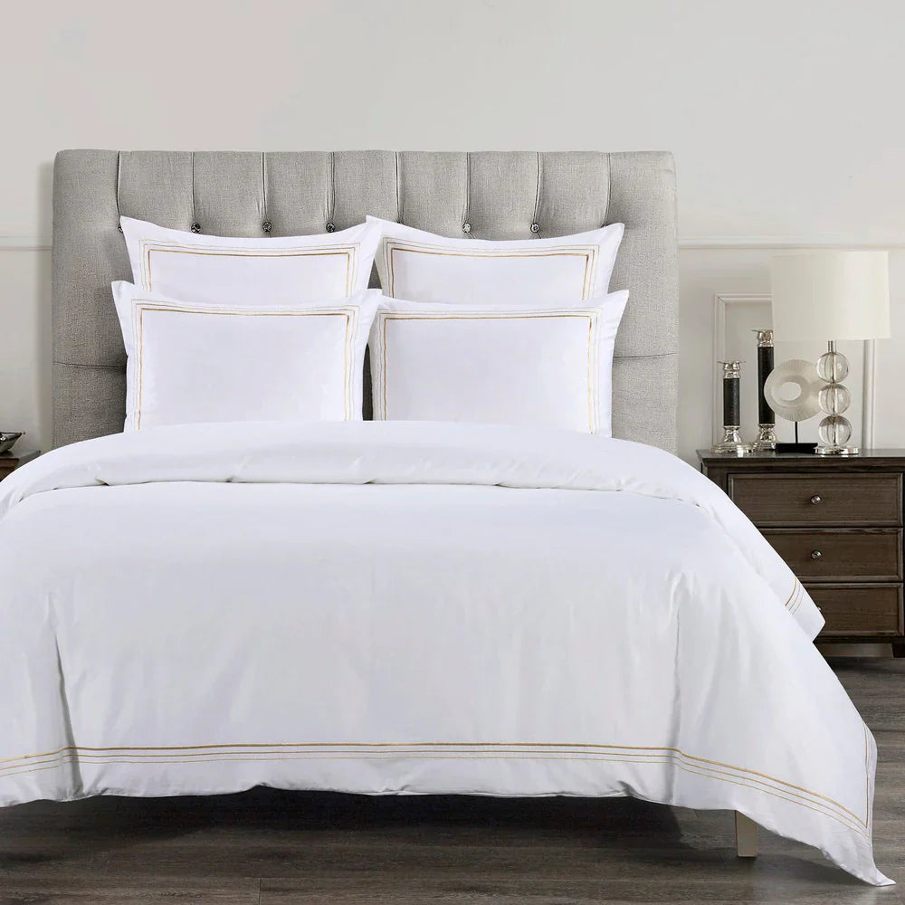 White bedding set with gold trim on a bed with a gray upholstered headboard.