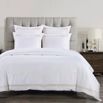 White bedding set with gold trim on a bed with a gray upholstered headboard.