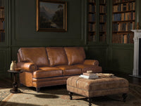 Brown leather sofa and ottoman in a room with bookshelves and a painting on the wall.