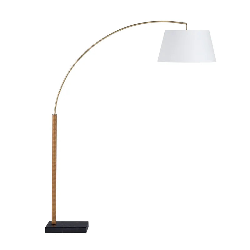 A brass floor lamp with a white lampshade, having a curved stand supported by a black base.