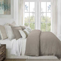Neatly made bed with beige and brown bedding in a bright room with large windows.