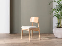 Wooden chair with a white cushion on a wooden floor against a gray wall with a plant in the corner.