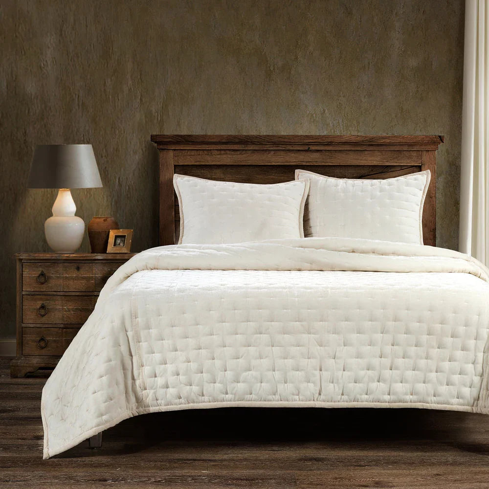 Bed with beige quilt and pillows in a bedroom setting with wooden nightstand and lamp.