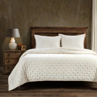 Bed with beige quilt and pillows in a bedroom setting with wooden nightstand and lamp.