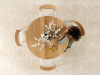 Person sitting at a round wooden table with a bowl of fruit and a vase of flowers, on a light textured floor.